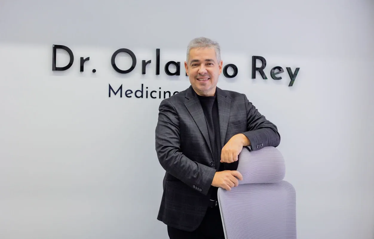 El Dr. Orlando Rey en su consultorio, apoyado en una silla frente al letrero con su nombre
